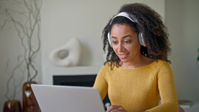 Satisfied African American woman with curly hair talking with friend through headphones. Cheerful lady using laptop during online communication in cosy home. Modern lifestyle. Digital technology.