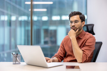 Serious businessman sitting in office working on laptop, hand on chin in thought. Professional environment with large windows in modern office setting. Phone on desk, indicating tech-savvy work.