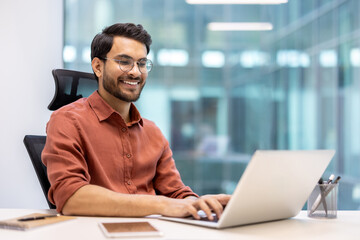 Smiling man wearing glasses and an orange shirt working on laptop in modern office. Phone and notebook on desk. Bright and professional workspace with glass walls.