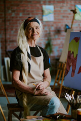 Vertical medium long portrait of cheerful senior female artist with long gray hair sitting on wooden stool next to easel with canvas on it