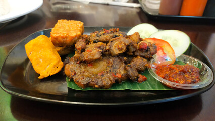 Spicy Grilled Oxtail with Tofu and Tempeh Served with Fresh Cucumber and Tomato Slices and Spicy Chili Sauce on Black Plate