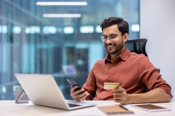 Young professional in a modern office using a phone and credit card for an online transaction. Displaying happiness and success in a business environment.