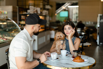 A woman with long hair with brown maltipoo and a bearded man in a cap are talking and eating croissants and drinking tea in cafe. Couple in love with little dog having breakfast in cafe.