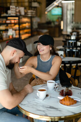 A woman with long hair with brown maltipoo and a bearded man in a cap are talking and eating croissants and drinking tea in cafe. Couple in love with little dog having breakfast in cafe.