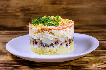 Plate with traditional russian salad Mimosa on a wooden table. Layered salad with potatoes, sardine, cheese, carrot, eggs and mayonnaise