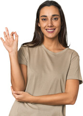 Hispanic young woman winks an eye and holds an okay gesture with hand.