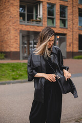 Elegant young woman looking in her black bag her phone or purse. Traveler style woman wear black leather shirt and bag on the street. Street style, fashion outfit, woman walk on the street