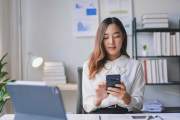 Young businesswoman is using a smartphone in a bright office, with a laptop on her desk and charts on the wall