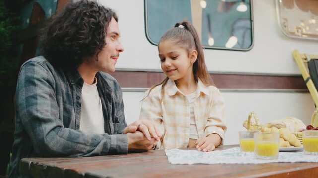 At a table next to their trailer, a father and daughter chat amiably while surrounded by orange juice, fresh fruits, and bread. The setting includes abundant greenery and home plants, enhancing the