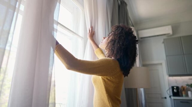 African American woman walking up to window and opening curtains in different sides. Curly female looking outdoor on street. Happily smiling while checking weather outside window. Peaceful day at home