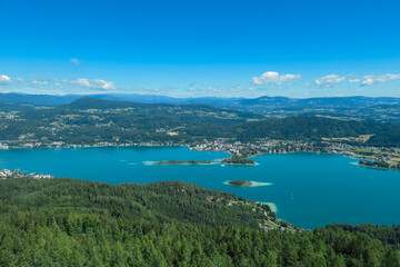 Fototapeta premium Aerial panoramic view of Lake Wörthersee seen from Pyramindenkogel in Carinthia, Austria. Serene turquoise waters, lush green forests and rolling hills of Austrian Alps. Charming lakeside village