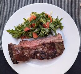 Piece of cooked meat, medium rare, and arugula tomato salad on white plate.