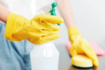 Woman in yellow rubber gloves holding a bottle of cleaning solution with a sponge on the table
