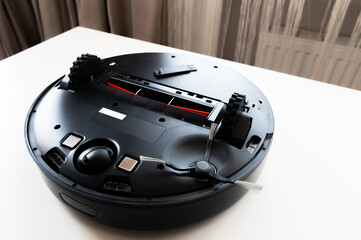 A black robot vacuum cleaner lies upside down on a white table. Cleaning and Maintenance of Home Cleaning Helper