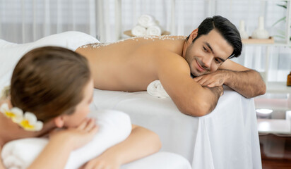 Blissful couple customer having exfoliation treatment in luxury spa salon with warmth candle light ambient. Salt scrub beauty treatment in health spa body scrub. Quiescent