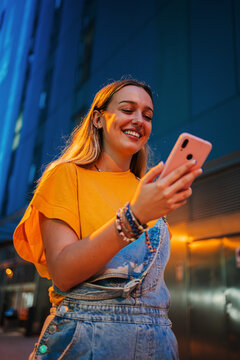 Vertical. one happy young girl using her smartphone to browse the internet while walking on the street at night. Female watching the social media with her cellphone. Girl typing on a mobile phone