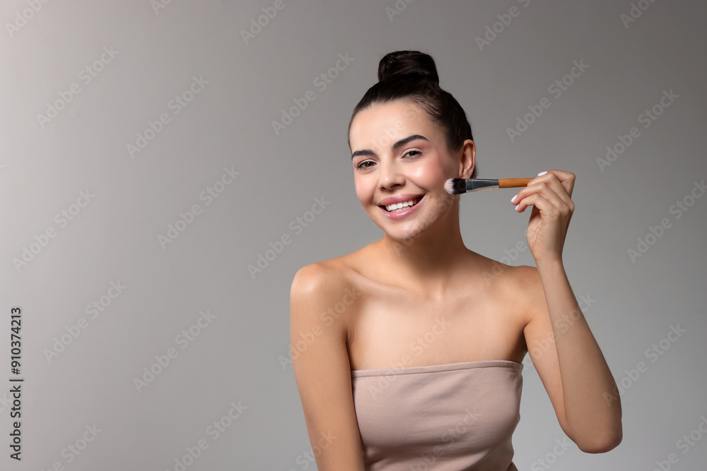 Wall mural Smiling woman applying makeup with brush on light grey background. Space for text