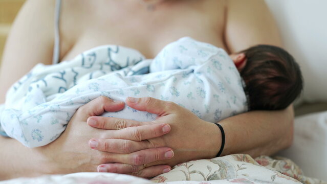 Close-up of mother hands holding newborn baby in arms while breastfeeding, maternal care of infant during first days of life