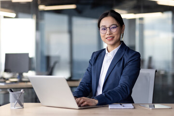 Fototapeta premium Professional Asian businesswoman working on a laptop at her desk in a modern office. She appears focused and confident, dressed in business attire, with a notepad and pen nearby.