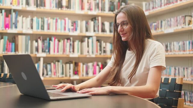 Caucasian positive female college student with a laptop watching a remote online seminar in the library, a live distance webinar or holding a virtual meetting in a campus, works on a business project.
