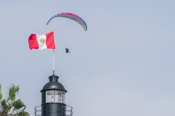 A colorful paraglider is flying over a lighthouse flying the Peruvian flag.
