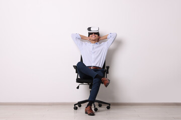 Happy young man with virtual reality headset sitting on chair near white wall