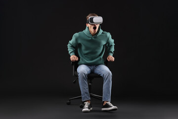 Emotional young man with virtual reality headset sitting on chair against black background