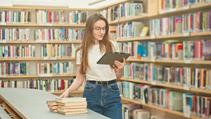 Caucasian woman wearing glasses with a tablet marking books in the library, working on a science project, doing homework for a tutor. Book selection, teacher prep, higher education, student e-learning