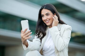 A young woman in a white blazer and white shirt smiles as she uses her smartphone and earphones in a modern urban environment. The woman is happy and relaxed, enjoying her time outdoors