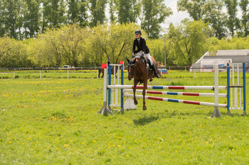 A young girl goes in for horse riding. A horse jumps over a barrier.