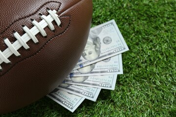 American football ball and dollar banknotes on green grass outdoors, closeup