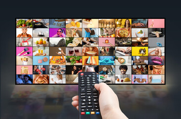 A persons hand holds a remote control and points it towards a large flat screen television. The TV screen displays a variety of colorful, diverse content in a grid format