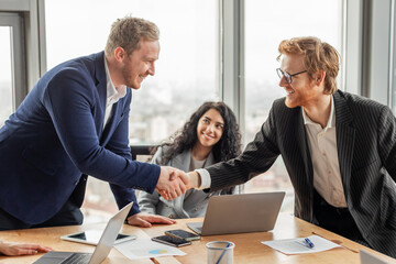 Two businessmen in suits are shaking hands in an office setting. They are both smiling and appear...