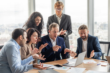 Fototapeta premium A group of business professionals gather around a laptop in a modern office setting, displaying excited reactions to the information on the screen. They are gathered around a large wooden table