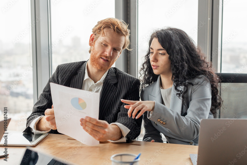 Aufkleber A man and a woman in business attire are reviewing a data graph during a meeting in a modern office. The man is seated at a table, holding the graph and looking at it intently