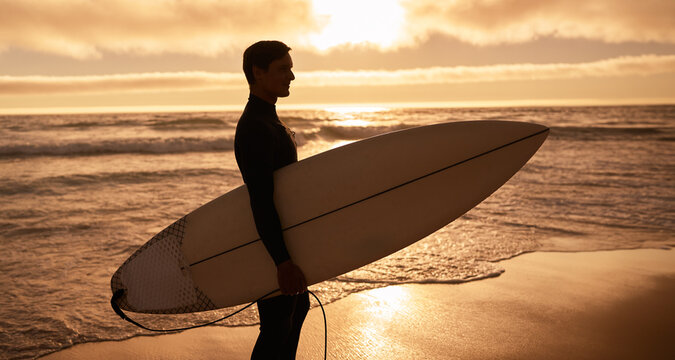 A surfer holding a surfboard while standing on the beach at sunset. The scene evokes feelings of adventure and serenity.