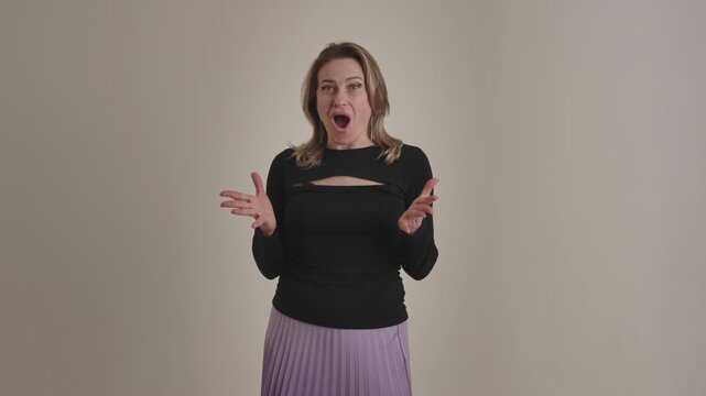 A young american or european excited and surprised blonde woman says wow, spreads hands on a beige studio background. An amazed and dumbfounded lady accepts unbelievable unexpected news, shows