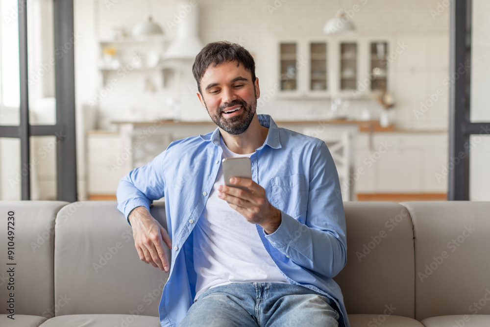 Poster a man with a beard sits on a light gray couch in a living room setting. he is wearing a white t-shir