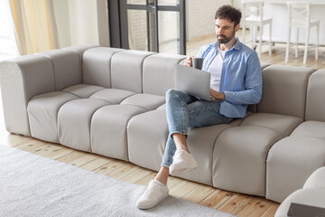 A man in a blue shirt and jeans sits on a light-colored sectional couch, working on a laptop computer while holding a cup of coffee in his other hand, copy space