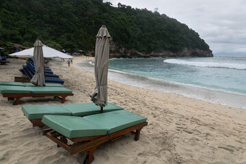 Row of empty deck chairs in cloudy weather on the Atuh beach on the island of Nusa Penida. Sandy beach, green islands,  sea surf and tropical nature around