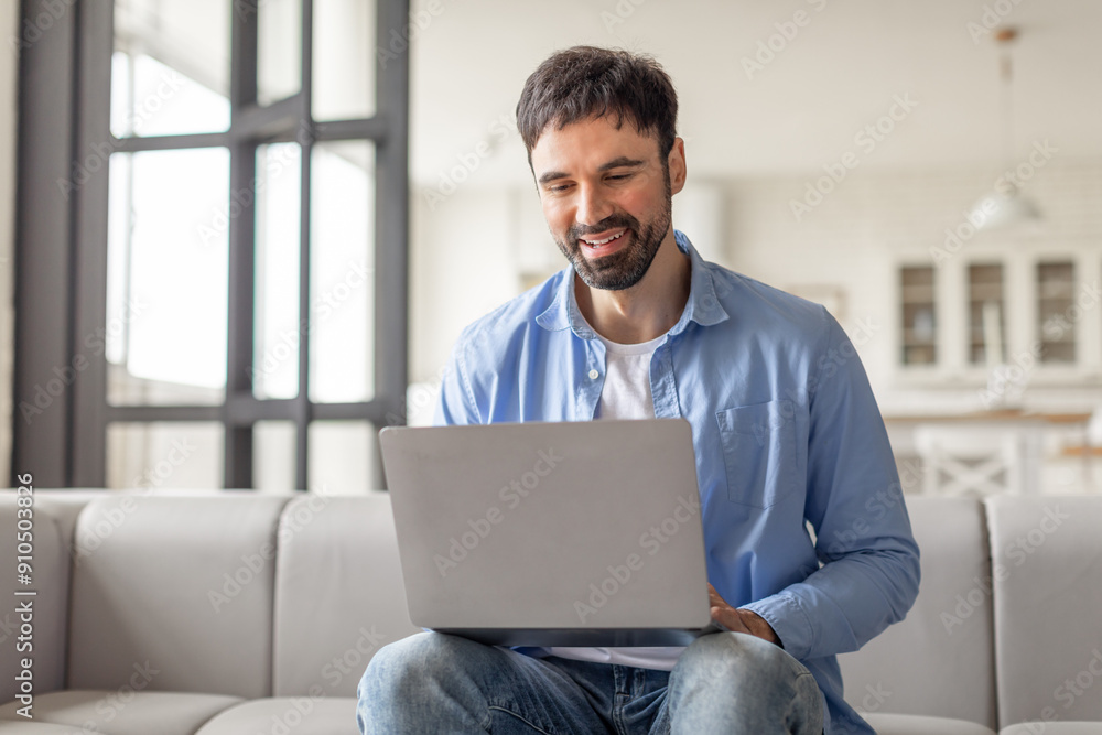 Poster A man sits on couch in a modern home, smiling as he uses a laptop computer. He is dressed casually in a blue shirt and jeans. The home is bright and airy, with large windows letting in natural light.