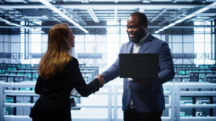 African american admin greeting inspector arriving in data center to supervise operations. IT expert in server hub workspace doing hand shake with supervisor monitoring performance, camera B