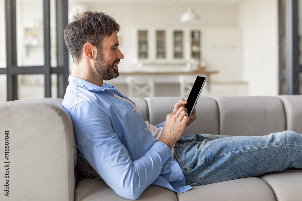 Poster A man is relaxing on a comfortable couch, using a tablet, in a modern home. He is sitting with his legs extended and leaning back against a pillow. He appears to be scrolling through content