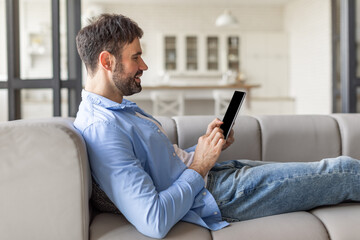 A man is relaxing on a comfortable couch, using a tablet, in a modern home. He is sitting with his legs extended and leaning back against a pillow. He appears to be scrolling through content