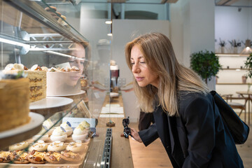 Unsmiling woman at display case with cakes, cookies, donuts, cupcakes, pastries chooses dessert for breakfast, lunch, dinner. Attentive businesswoman thoughtfully buys purchase snack in a coffee house