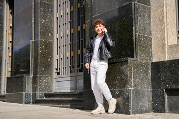 A man in a black leather jacket and white pants is talking on her cell phone. She is standing in front of a building