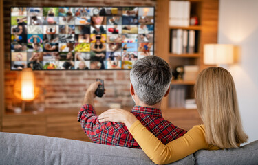 A couple is relaxing on a couch in their living room. The man is holding a remote control and is...