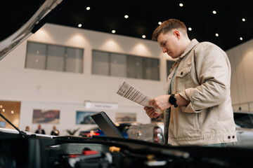 Shooting from below of thoughtful male customer holding papers, checking vehicle characteristics during choosing car in modern dealership center. Concept of choosing and buying new auto at showroom.