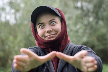 A funny girl in a cap and hood stretching out her arms with a strange smile and look in nature