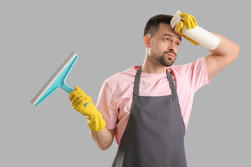 Tired male janitor with squeegee and detergent on grey background